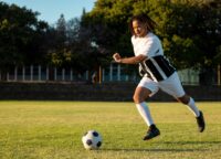 Male soccer player running toward ball on sunlit field, with striped jersey, cleats, copy space. Athlete, sportswear, kicking, outdoor, grass, dynamic, determination