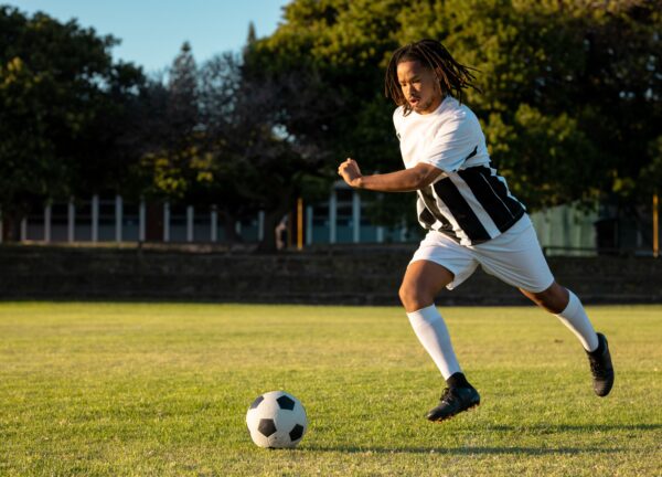 Male soccer player running toward ball on sunlit field, with striped jersey, cleats, copy space. Athlete, sportswear, kicking, outdoor, grass, dynamic, determination