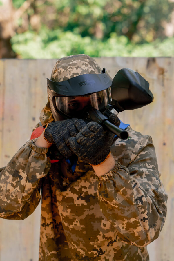 close up in the wild near tree structures a player in a pintball uniform holds a pistol with a red balloon team game camouflage clothing